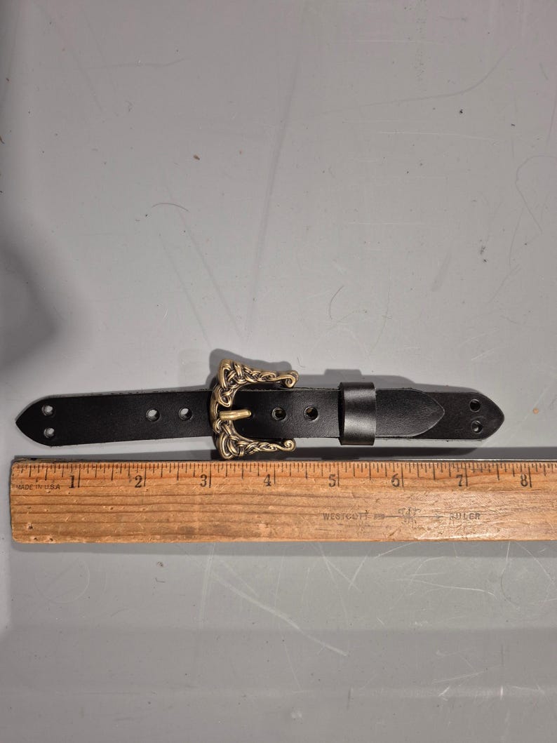 May include: A black leather belt with an ornate gold-colored buckle. The belt has a rectangular shape with a series of holes for adjustment. The buckle features a detailed, vintage design. The belt measures approximately 8 inches in length.