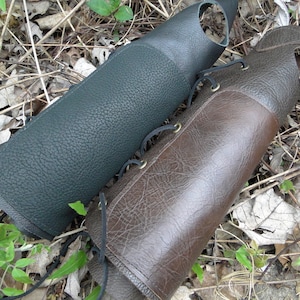 May include: Two leather arm guards, one dark green and one brown, with laces. The guards are lying on a bed of dry leaves and twigs.