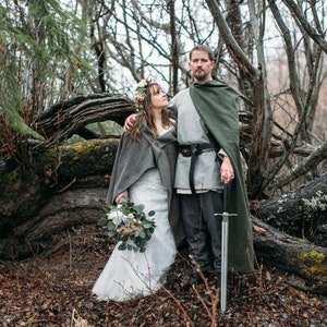 May include: A couple in a forest setting, the woman wearing a white wedding dress and a gray shawl, the man wearing a gray tunic and a green cloak, holding a sword.
