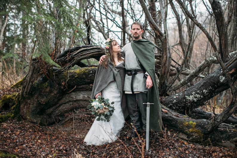 May include: A couple in a forest setting, the woman wearing a white wedding dress and a gray shawl, the man wearing a gray tunic and a green cloak, holding a sword.