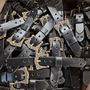 May include: A collection of black leather belt straps with ornate, antique brass-colored buckles. The buckles have a decorative, vintage design. The straps are piled together, showing the details of the leather and metal.