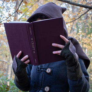 May include: A person wearing a brown hooded cloak and fingerless gloves is reading a book titled "CRAFT" in a forest setting.