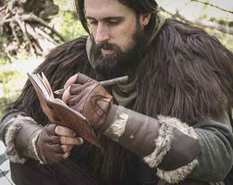 May include: A person wearing a brown fur-lined cloak and leather gloves writes in a brown leather journal with a twig.