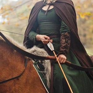 May include: A woman in a green dress and brown cloak is riding a horse while holding a bow and arrow. She is wearing leather arm guards and a belt with a buckle. The horse is brown and has a white mane and tail.