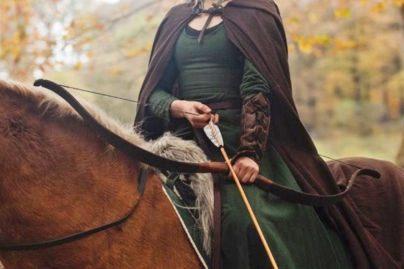 May include: A woman in a green dress and brown cloak is riding a horse while holding a bow and arrow. She is wearing leather arm guards and a belt with a buckle. The horse is brown and has a white mane and tail.