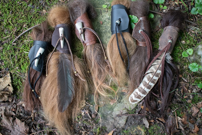 May include: Five brown leather hair wraps with feathers and bone accents. The wraps are on a bed of green moss and brown leaves.