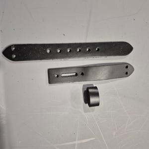 May include: Three pieces of dark gray leather, likely for a belt or strap. One long, rectangular piece has multiple holes. Another shorter piece has a rectangular slot. A small, cylindrical piece is also present.