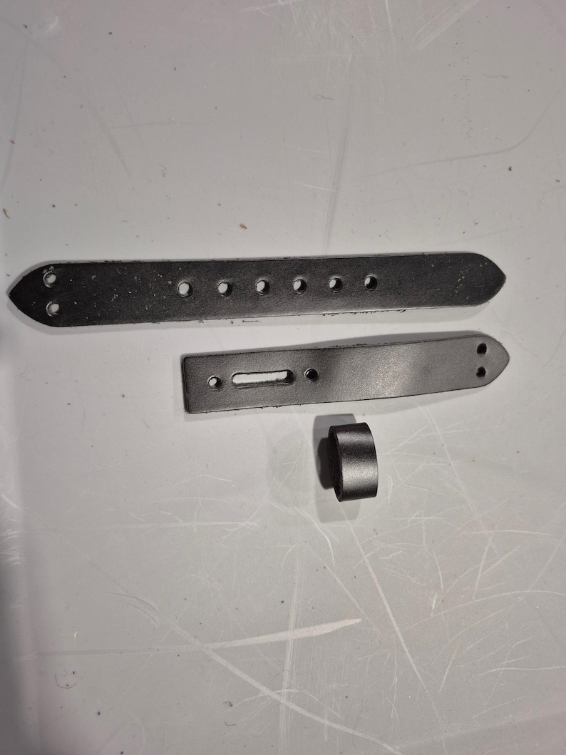 May include: Three pieces of dark gray leather, likely for a belt or strap. One long, rectangular piece has multiple holes. Another shorter piece has a rectangular slot. A small, cylindrical piece is also present.