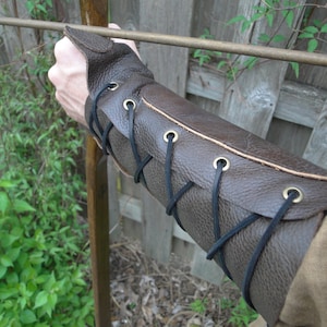 May include: Brown leather arm guard with black laces and brass eyelets. The guard is worn on the left arm and is secured with laces.