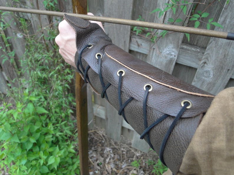 May include: Brown leather arm guard with black laces and brass eyelets. The guard is worn on the left arm and is secured with laces.