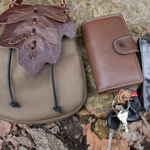 May include: A brown leather wallet, a small brown leather bag with a leaf design, a black cell phone, and a set of keys with a key fob on a brown and gray background.