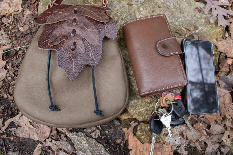 May include: A brown leather wallet, a small brown leather bag with a leaf design, a black cell phone, and a set of keys with a key fob on a brown and gray background.