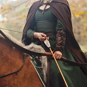May include: A woman in a green dress and brown cloak is riding a horse while holding a bow and arrow. She is wearing leather arm guards and a belt with a buckle. The horse is brown and has a white mane and tail.