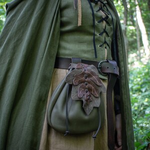 May include: A green leather pouch with a brown leaf design hangs from a brown leather belt. The pouch is worn by a person in a forest setting.