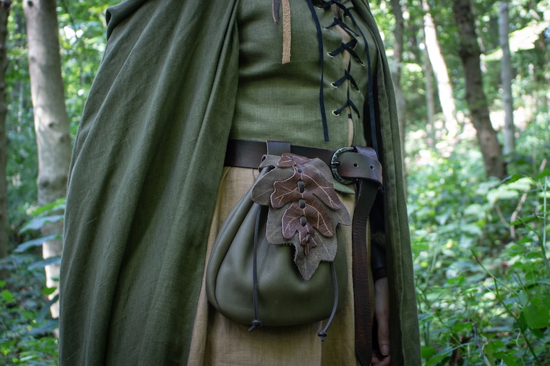 May include: A green leather pouch with a brown leaf design hangs from a brown leather belt. The pouch is worn by a person in a forest setting.