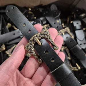 May include: A black leather belt with an ornate, antique brass-colored buckle. The buckle features a detailed, interwoven design. The belt has three holes for adjustment and a loop to secure the end. Other belts are visible in the background.