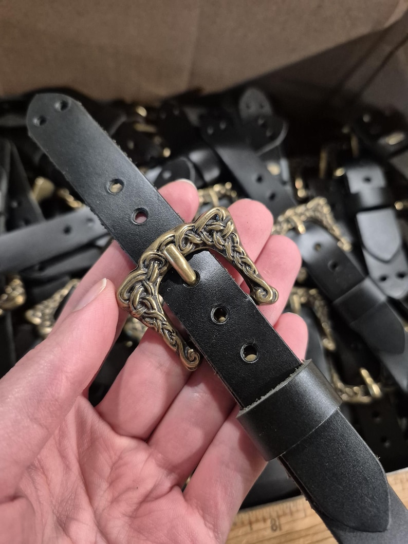 May include: A black leather belt with an ornate, antique brass-colored buckle. The buckle features a detailed, interwoven design. The belt has three holes for adjustment and a loop to secure the end. Other belts are visible in the background.