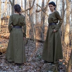 May include: A long, olive green linen dress with a brown leather belt. The dress has a simple, loose fit and is worn by a person standing in a forest setting.