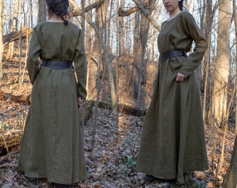 Puede incluir: Un vestido largo de lino verde oliva con un cinturón de cuero marrón. El vestido tiene un corte sencillo y holgado y lo lleva puesto una persona de pie en un entorno boscoso.