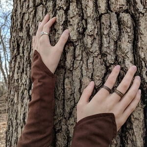 May include: Two hands wearing brown fabric sleeves reach out to touch a rough, brown tree bark. One hand is wearing a silver ring with a decorative design. The other hand is wearing two thin, brown rings.