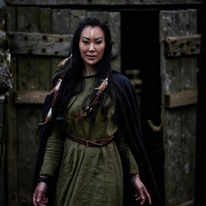 May include: A woman with long black hair and a white stripe painted on her forehead wears a long, dark green linen dress with a brown leather belt. She is wearing a black cloak and has feathers in her hair.