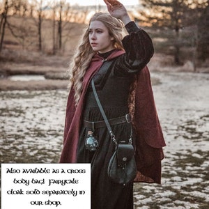 May include: A woman wearing a burgundy cloak with a black velvet bodice and a black leather belt. She is carrying a black leather bag with a silver chain and a small glass bottle. The text "Also available as a cross body bag! Fairycale cloak sold separately in our shop." is visible in the image.