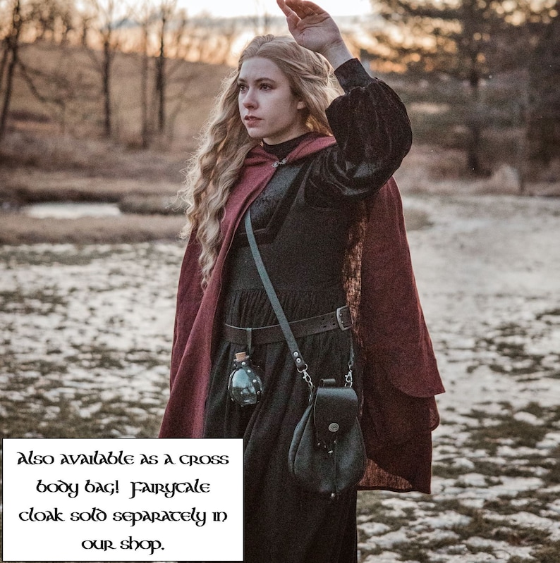May include: A woman wearing a burgundy cloak with a black velvet bodice and a black leather belt. She is carrying a black leather bag with a silver chain and a small glass bottle. The text "Also available as a cross body bag! Fairycale cloak sold separately in our shop." is visible in the image.