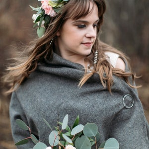 May include: A woman wearing a gray shawl and a flower crown with pink and white flowers. She is holding a bouquet of white and pink roses with eucalyptus leaves.