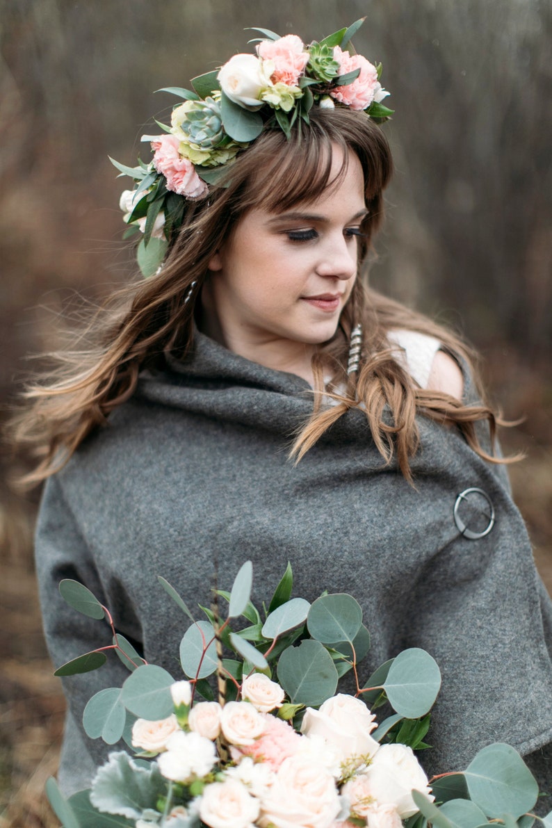 May include: A woman wearing a gray shawl and a flower crown with pink and white flowers. She is holding a bouquet of white and pink roses with eucalyptus leaves.