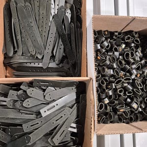 May include: A collection of dark gray leather straps with multiple holes and rounded ends, neatly arranged in cardboard boxes. The image also shows a box filled with black, circular leather pieces.