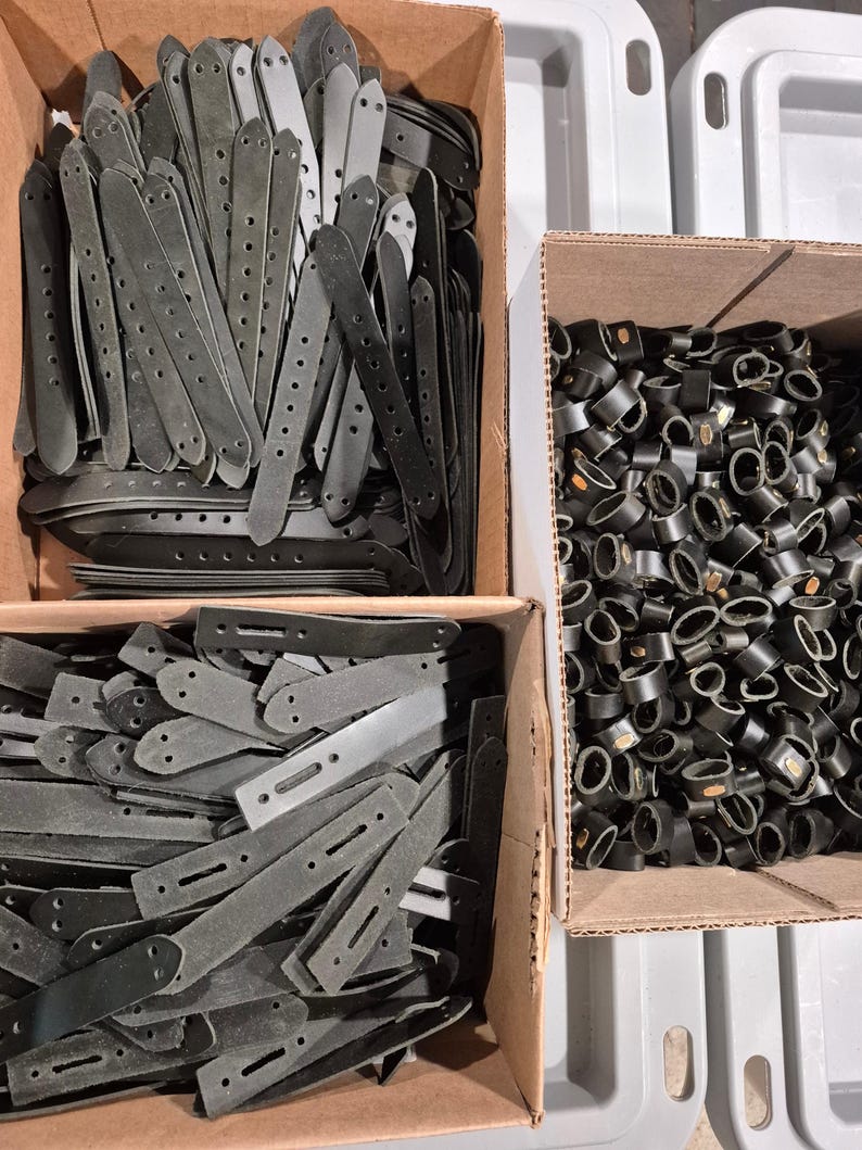 May include: A collection of dark gray leather straps with multiple holes and rounded ends, neatly arranged in cardboard boxes. The image also shows a box filled with black, circular leather pieces.