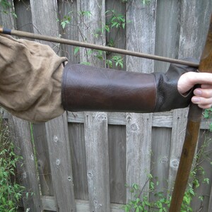 May include: A brown leather arm guard with a wooden bow and arrow. The arm guard is worn on the left arm and is secured with a strap. The bow is held in the right hand.