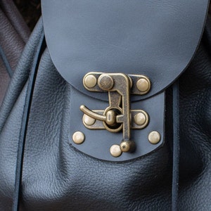May include: A black leather backpack with a brass buckle and metal accents. The backpack has a flap closure and a drawstring top.