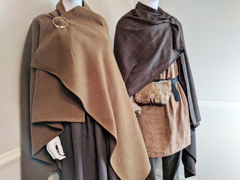 May include: Two mannequins wearing brown cloaks. The cloak on the left is a solid brown color and is fastened with a circular metal clasp. The cloak on the right is a plaid pattern with brown, black, and red colors. The mannequin on the right is also wearing a brown tunic and a brown leather belt with a fur pouch.