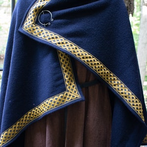May include: A dark blue wool cloak with a gold Celtic knot trim. The cloak is fastened with a silver ring brooch.