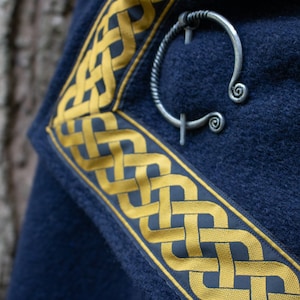 May include: A dark blue fabric with a gold Celtic knot border. A silver Celtic knot brooch is pinned to the fabric.