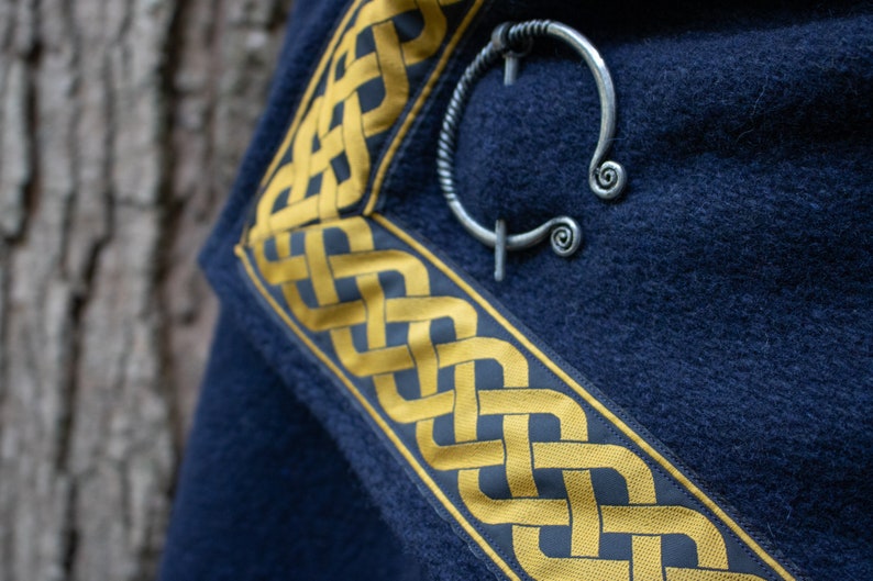 May include: A dark blue fabric with a gold Celtic knot border. A silver Celtic knot brooch is pinned to the fabric.