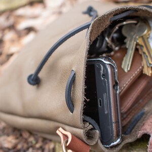 May include: A brown leather pouch with a drawstring closure. The pouch is open and contains a smartphone, keys, and a wallet.