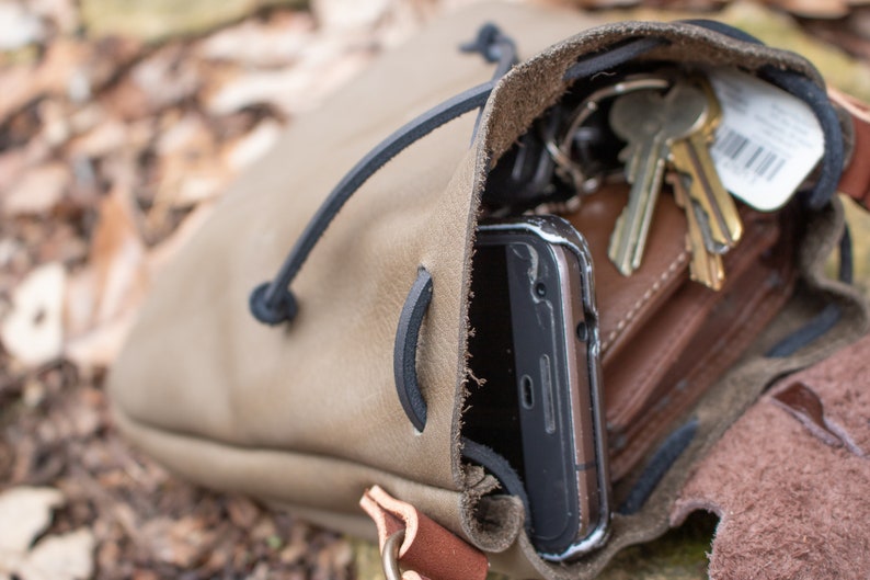 May include: A brown leather pouch with a drawstring closure. The pouch is open and contains a smartphone, keys, and a wallet.