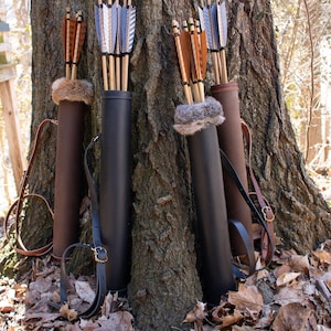 May include: Four brown leather arrow quivers with fur lining, each holding a bundle of arrows with different fletching, leaning against a tree trunk.