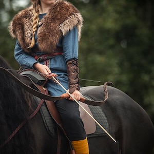May include: A woman in a blue tunic with a fur collar rides a black horse. She holds a bow and arrow in her hands.