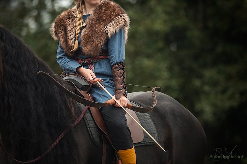 May include: A woman in a blue tunic with a fur collar rides a black horse. She holds a bow and arrow in her hands.
