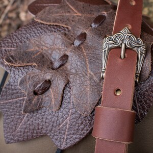 May include: A brown leather bag with a leaf-shaped flap and a brown leather strap with a silver buckle. The buckle has a Celtic knot design.
