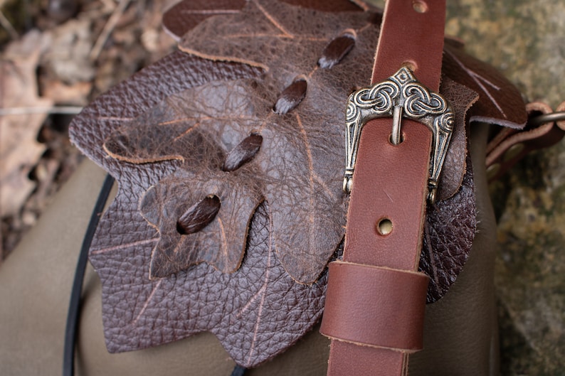 May include: A brown leather bag with a leaf-shaped flap and a brown leather strap with a silver buckle. The buckle has a Celtic knot design.