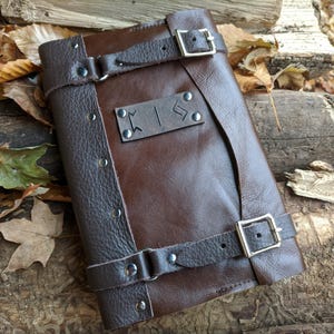 May include: A brown leather journal with dark brown detailing and straps fastened with silver buckles. The journal has a leather tag with etched symbols. The cover is a rich, warm brown, and it's secured with two leather straps.