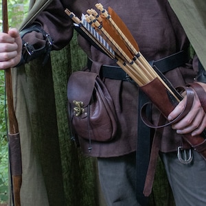 May include: A person wearing a brown tunic and a green hooded cloak is holding a quiver of arrows. The quiver is made of brown leather and has a strap that goes around the person's waist. The person is also wearing a brown leather belt with a metal buckle.