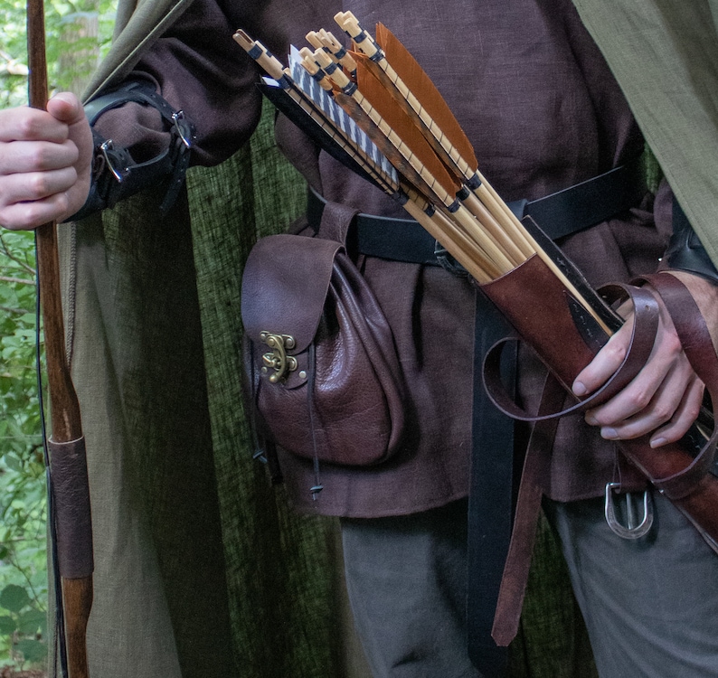 May include: A person wearing a brown tunic and a green hooded cloak is holding a quiver of arrows. The quiver is made of brown leather and has a strap that goes around the person's waist. The person is also wearing a brown leather belt with a metal buckle.