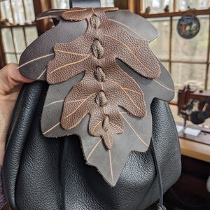 May include: A black leather drawstring bag with a brown leather leaf design. The bag has a strap and a drawstring closure.