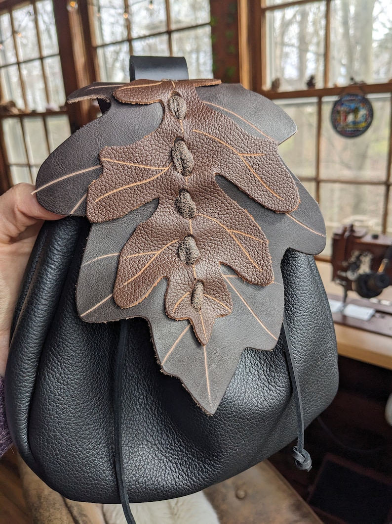 May include: A black leather drawstring bag with a brown leather leaf design. The bag has a strap and a drawstring closure.