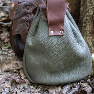 May include: A gray leather pouch with a brown leather strap and metal rivets. The pouch is sitting on a bed of brown leaves.